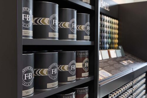 Display of Farrow & Ball paint cans on shelves next to a paint color sample wall in a store.