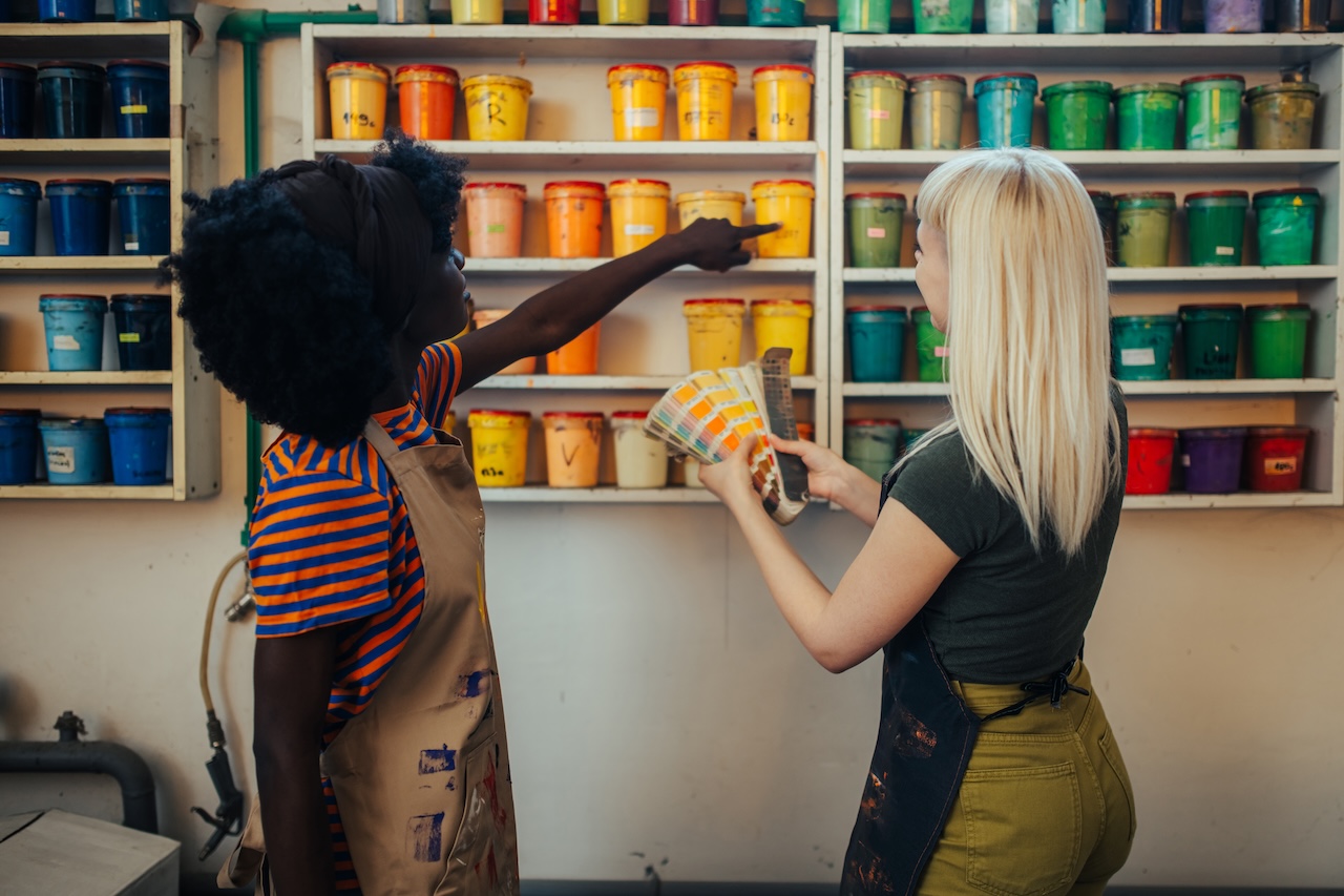 Two women with paint swatches selecting paint colors from a shelf filled with paint buckets